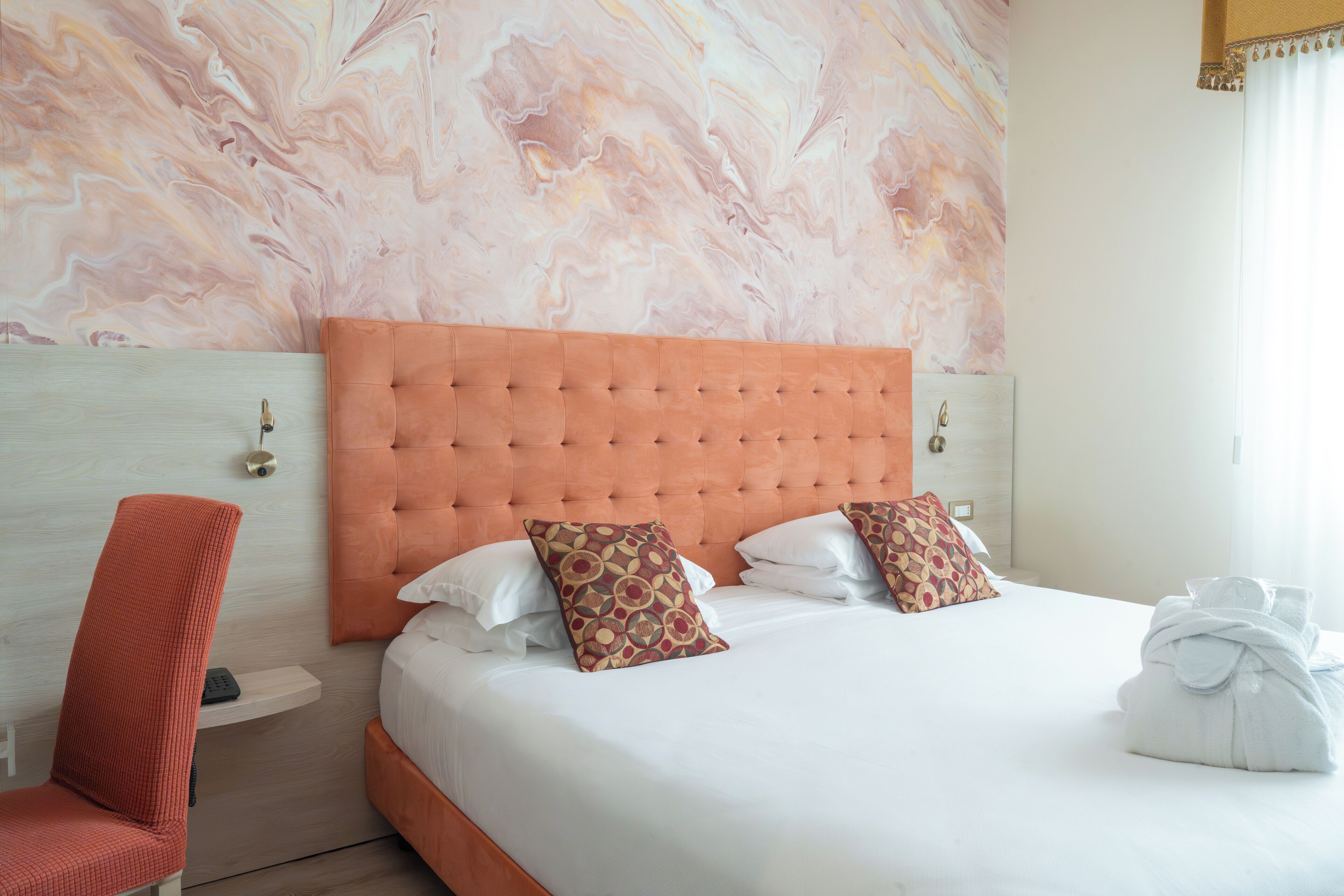 A hotel room with a marble-patterned wall, a tufted orange headboard, two patterned pillows, a white bedspread, and a neatly folded towel on the bed.