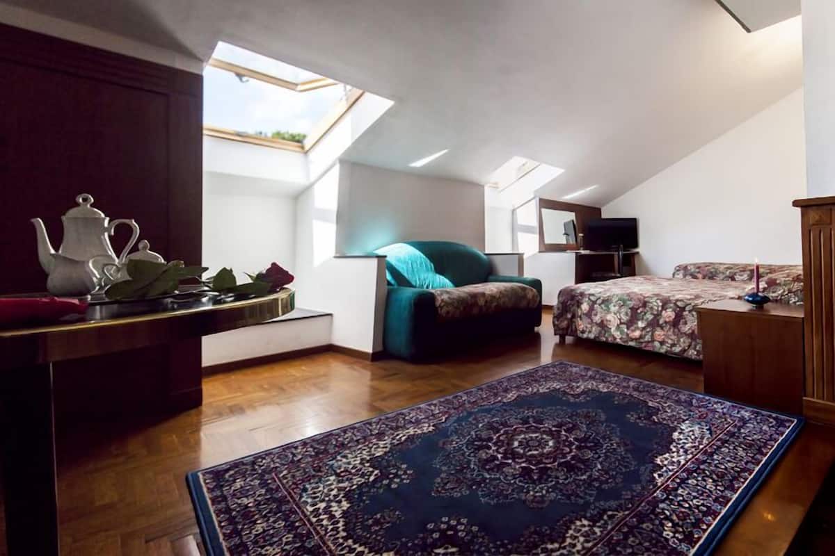 A compact, well-lit room with a sloped ceiling, a skylight, a bed, a sofa, a small table with a teapot, and a patterned rug.