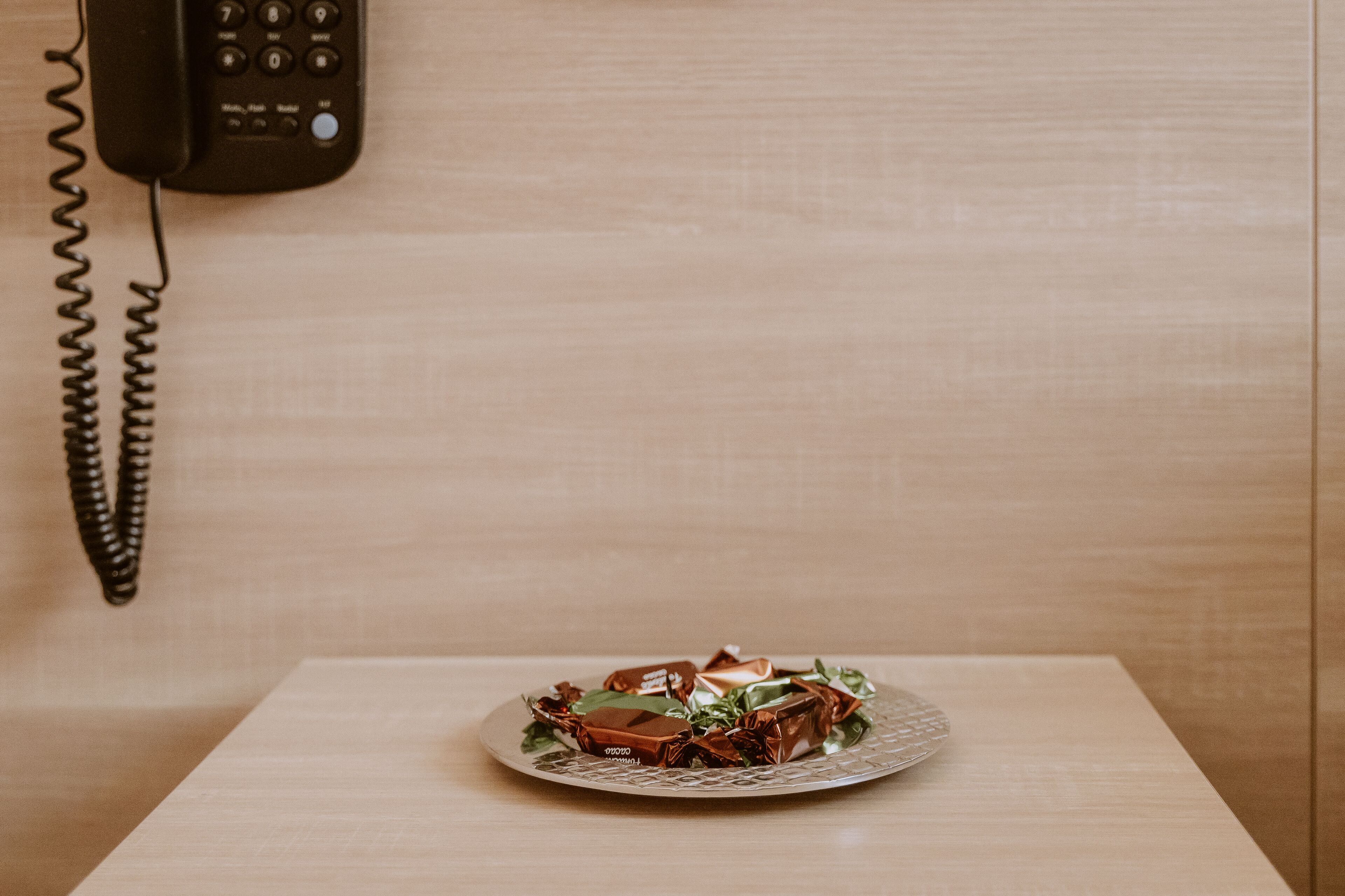 A wooden table with a plate of wrapped chocolates and a telephone on the wall.