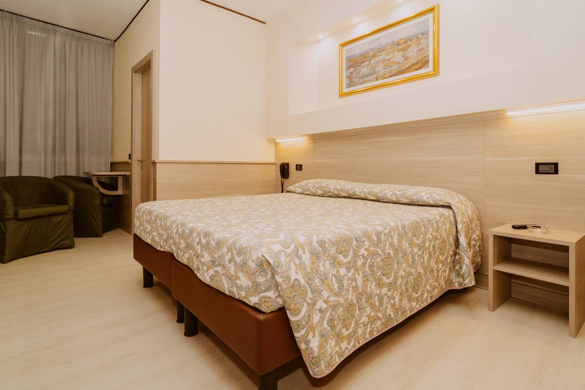 A hotel room with a bed, a chair, a bedside table, and a painting on the wall.