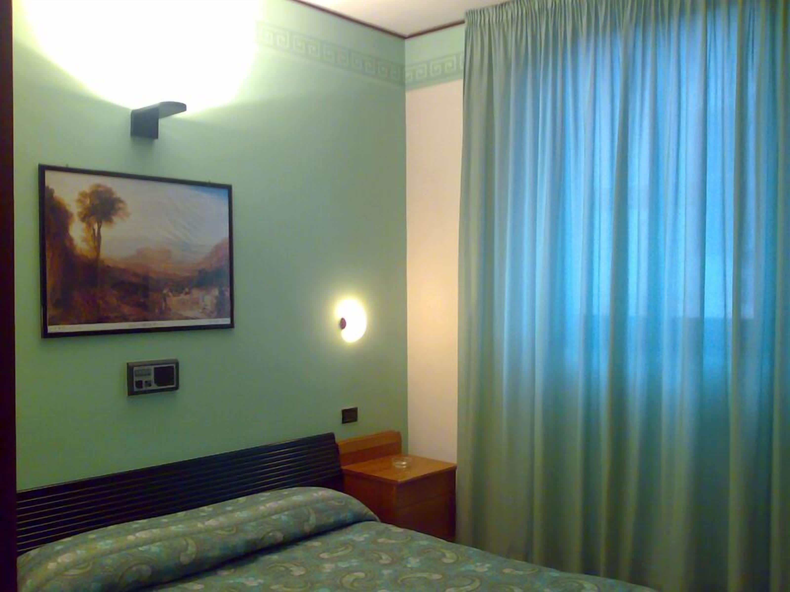 A hotel room with a bed, a nightstand, a painting, a wall-mounted light, and a window with curtains.