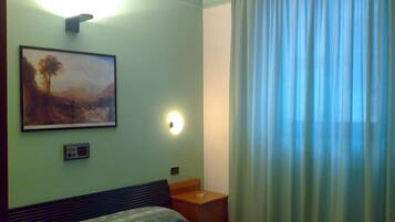 A hotel room with a bed, a nightstand, a painting, a wall-mounted light, and a window with curtains.