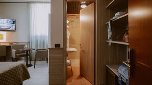 A hotel room with a bed, a desk with a chair, a small table with a lamp, a bathroom with a sink and shower, and a wardrobe with shelves. - Hotel Valentino (Orvieto)