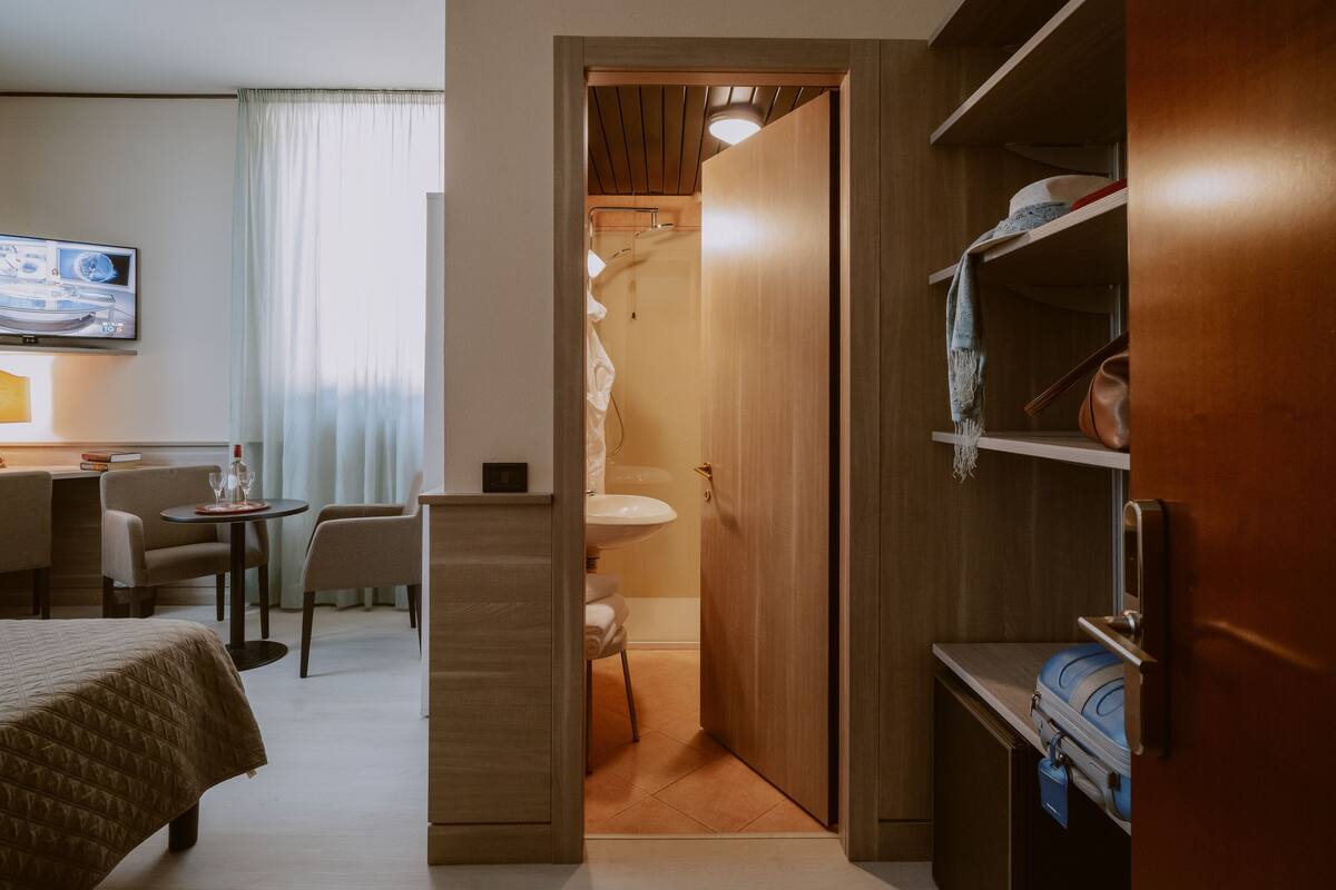 A hotel room with a bed, a desk with a chair, a small table with a lamp, a bathroom with a sink and shower, and a wardrobe with shelves.