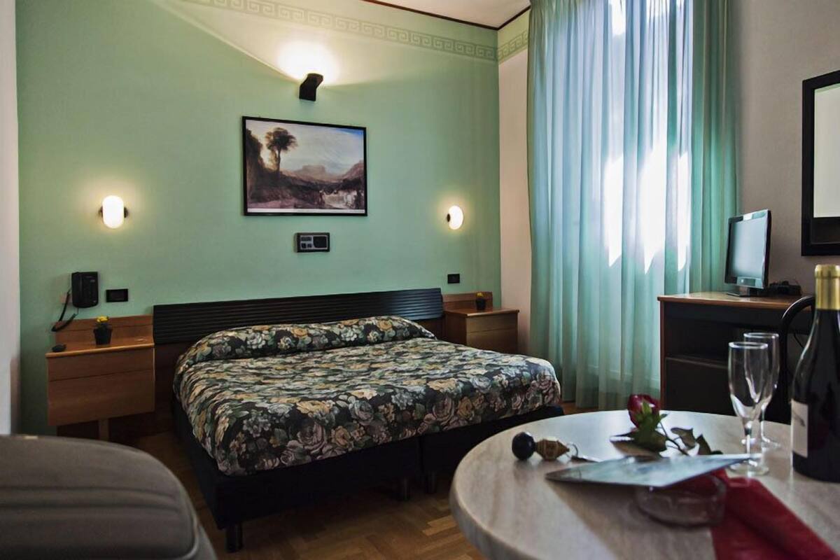 A hotel room with a bed, bedside tables, a desk with a chair, a television, and a dining table with a wine glass and a rose.