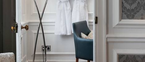 A room with a bed, a chair, a lamp, and a door leading to a bathroom with two white bathrobes hanging on a rack.