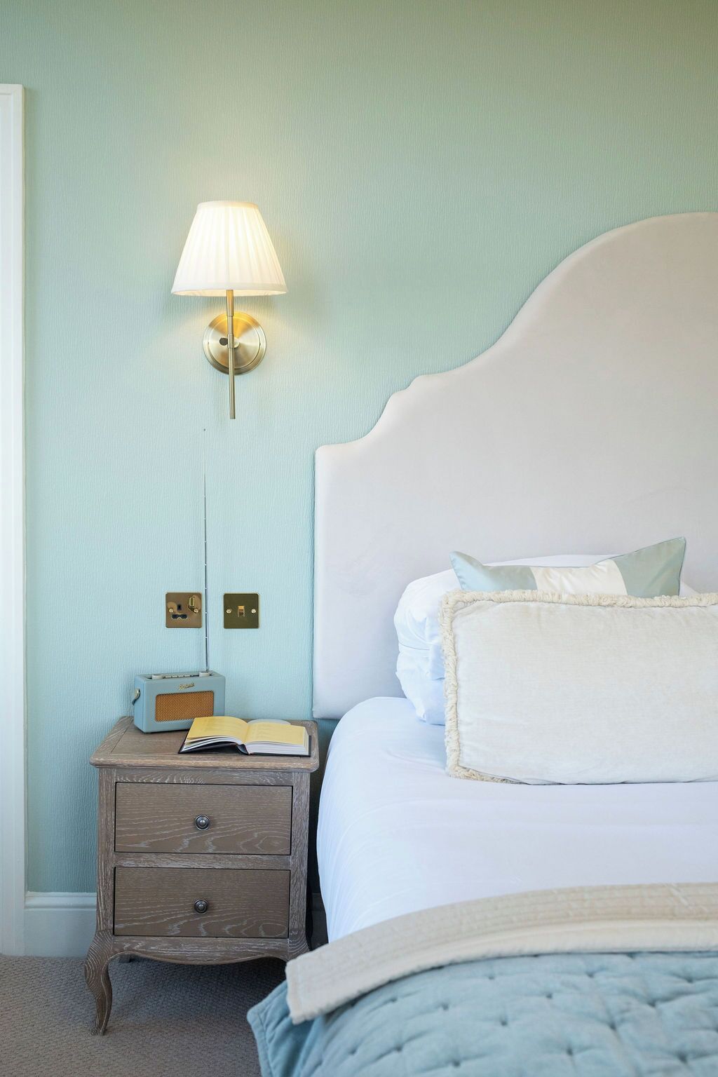 Quarto com cabeceira branca, luminária de cabeceira, rádio e um livro sobre uma mesinha de madeira.