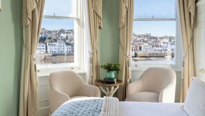 A bedroom with a bed, two chairs, a small table with a book and a vase, and a view of a town through large windows. - The Royal Hotel (Bideford)