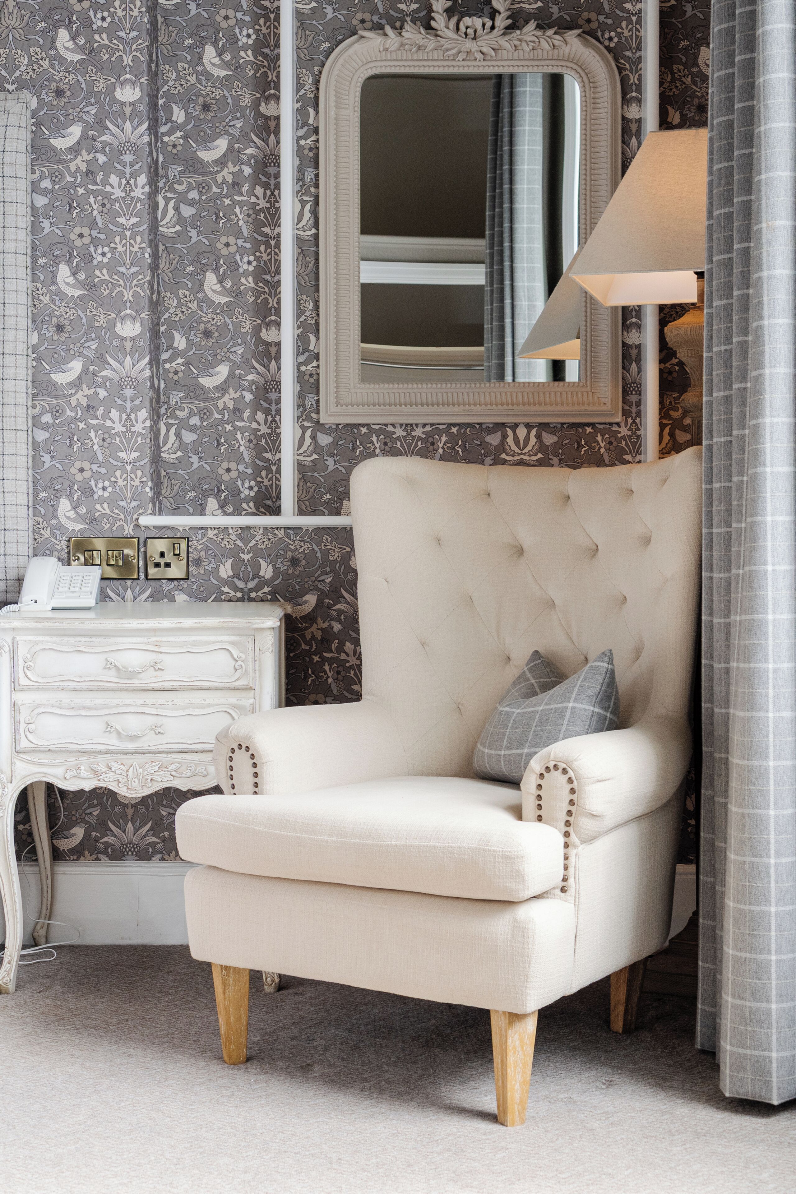 Een kamer met een gestoffeerde fauteuil, een witte kaptafel met een telefoon en fotolijstjes, en een spiegel boven een raam met gordijnen.