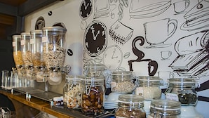 Un espace buffet proposant divers aliments présentés dans des bocaux et des bols, une machine à café et un mur décoré de dessins de montres et d’ustensiles de cuisine.