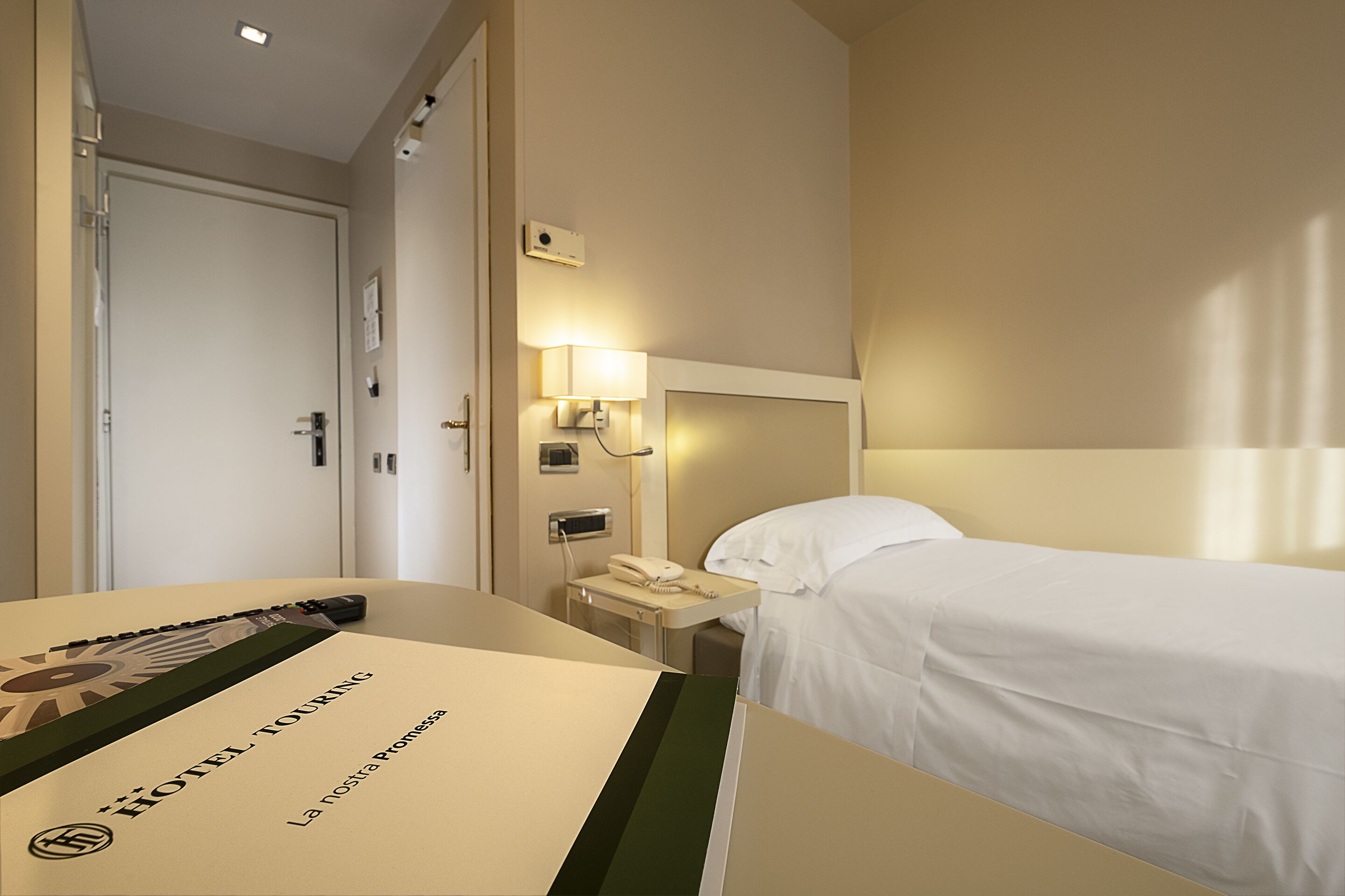 A hotel room with a bed, a bedside table with a lamp, a phone, and a door.