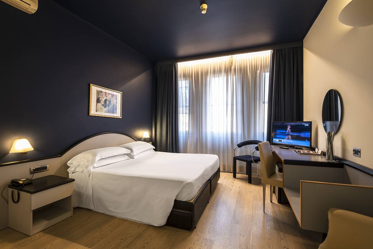 A hotel room with a bed, bedside table, desk, chair, and a TV.