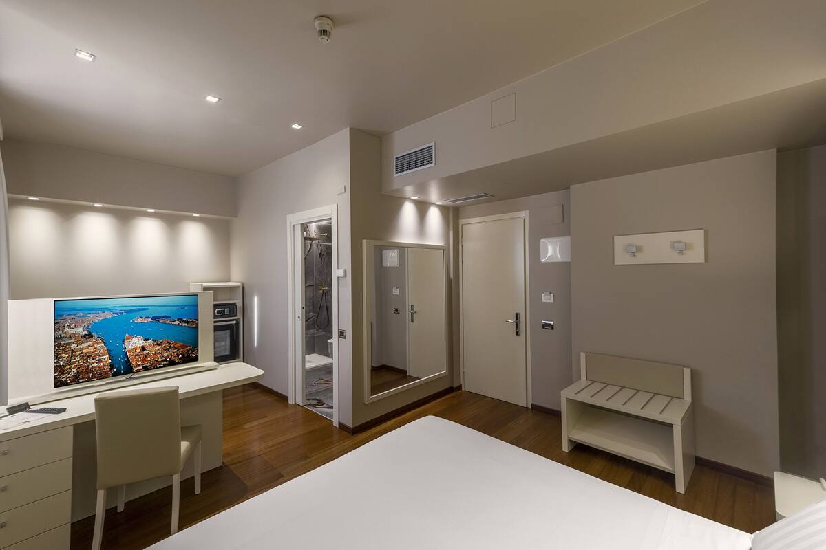 A modern bedroom with a large bed, a desk with a TV, a mirror, and a small bench.
