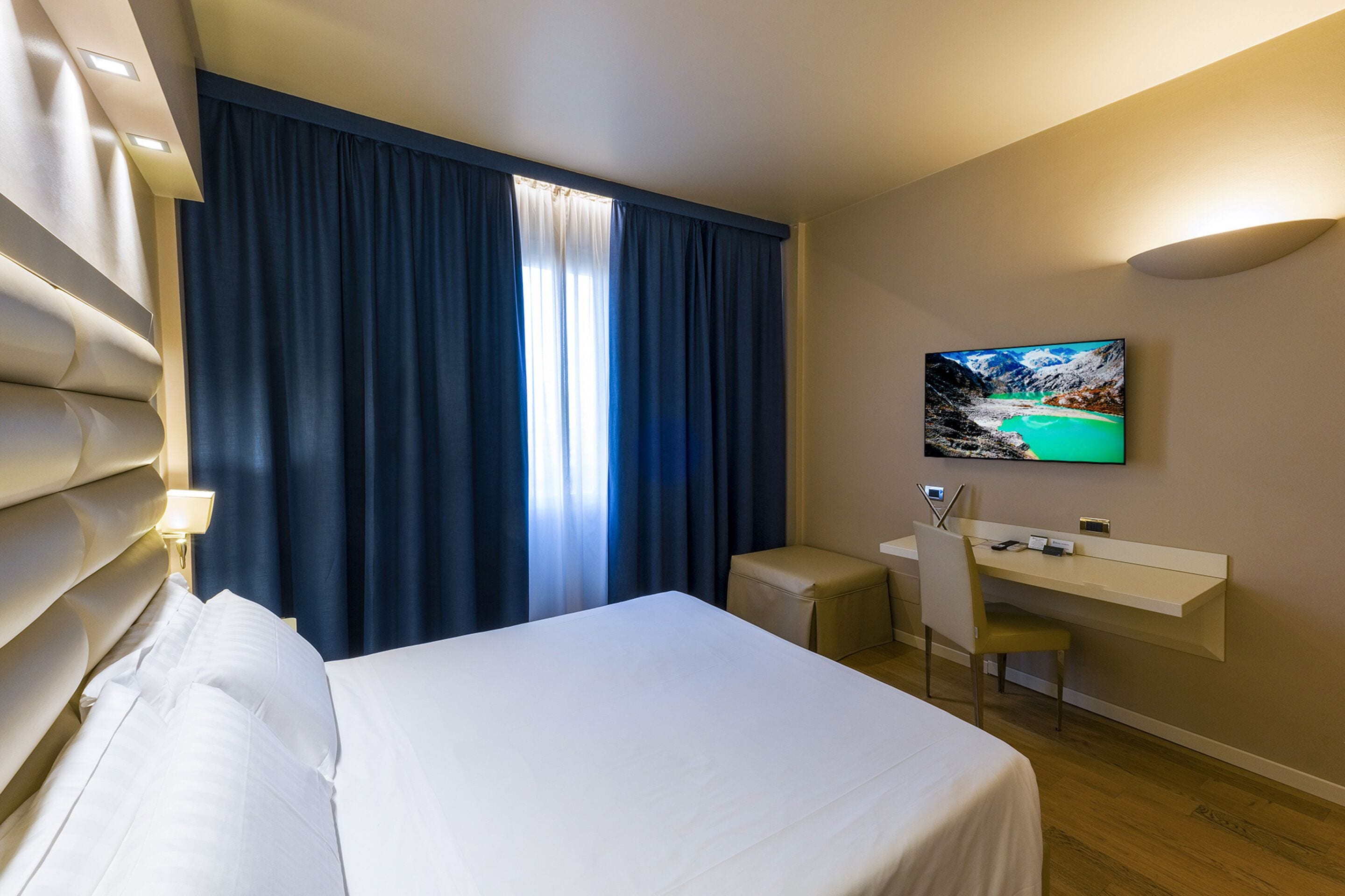 A hotel room with a bed, a desk, a chair, a TV, and a window with curtains.
