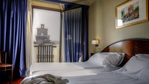 A hotel room with a bed, a chair, a balcony with a view of a building, and a framed picture on the wall.