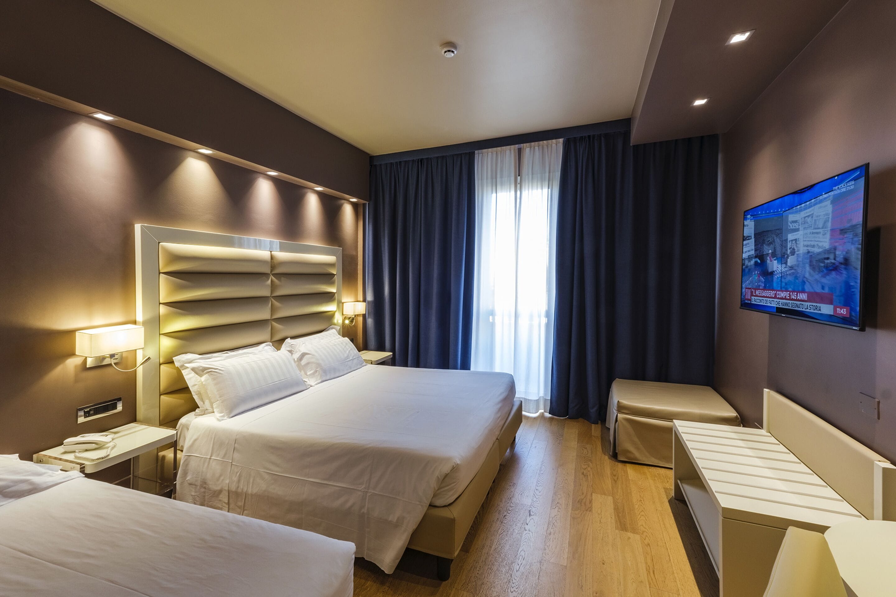 Habitación de hotel con dos camas, un televisor de pantalla plana montado en la pared y una ventana con cortinas.