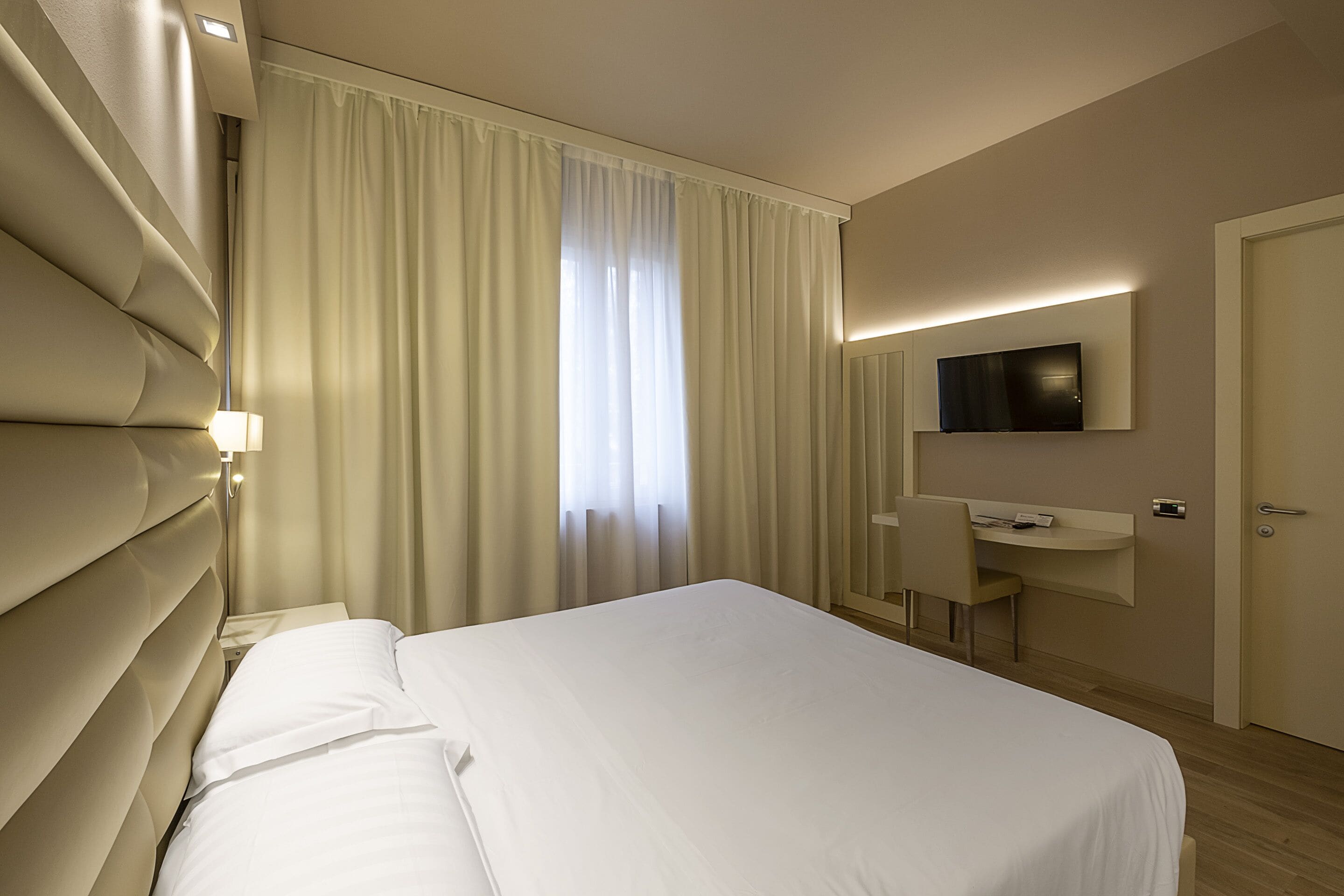 A hotel room with a bed, a desk, a chair, a television mounted on the wall, and a door.