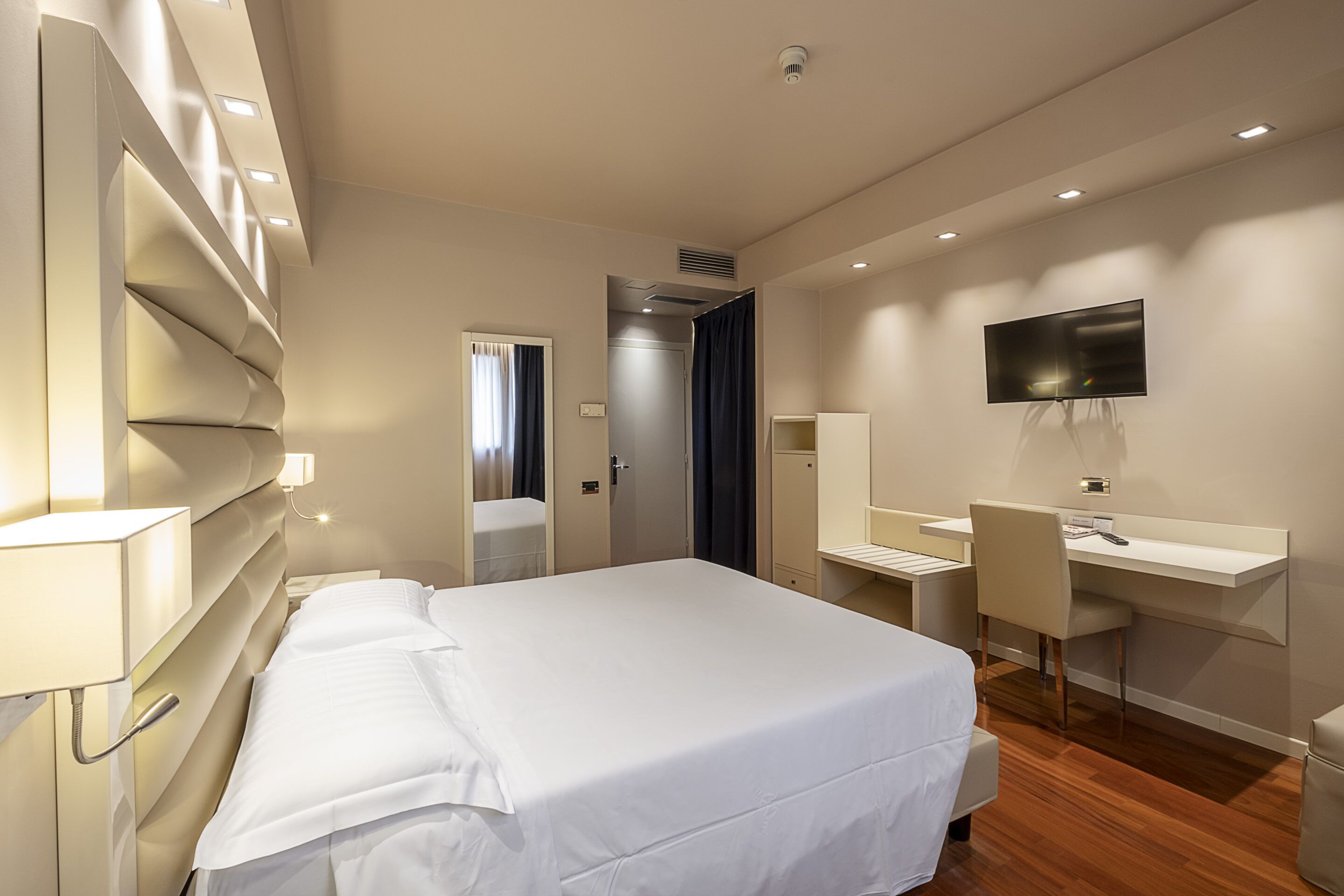 A modern hotel room with a large bed, a desk, a chair, a TV, and a mirror.