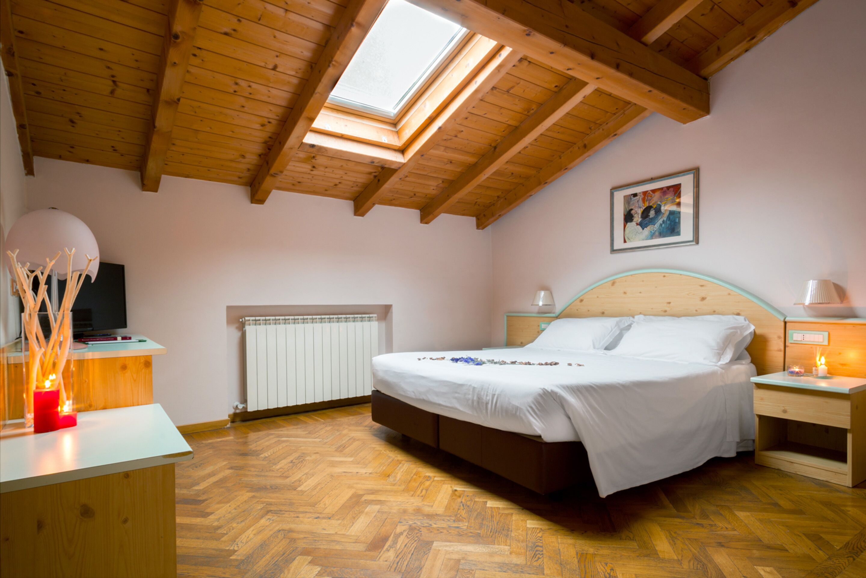 A bedroom with a wooden ceiling, a skylight, a bed, a bedside table with a lamp, and a radiator.