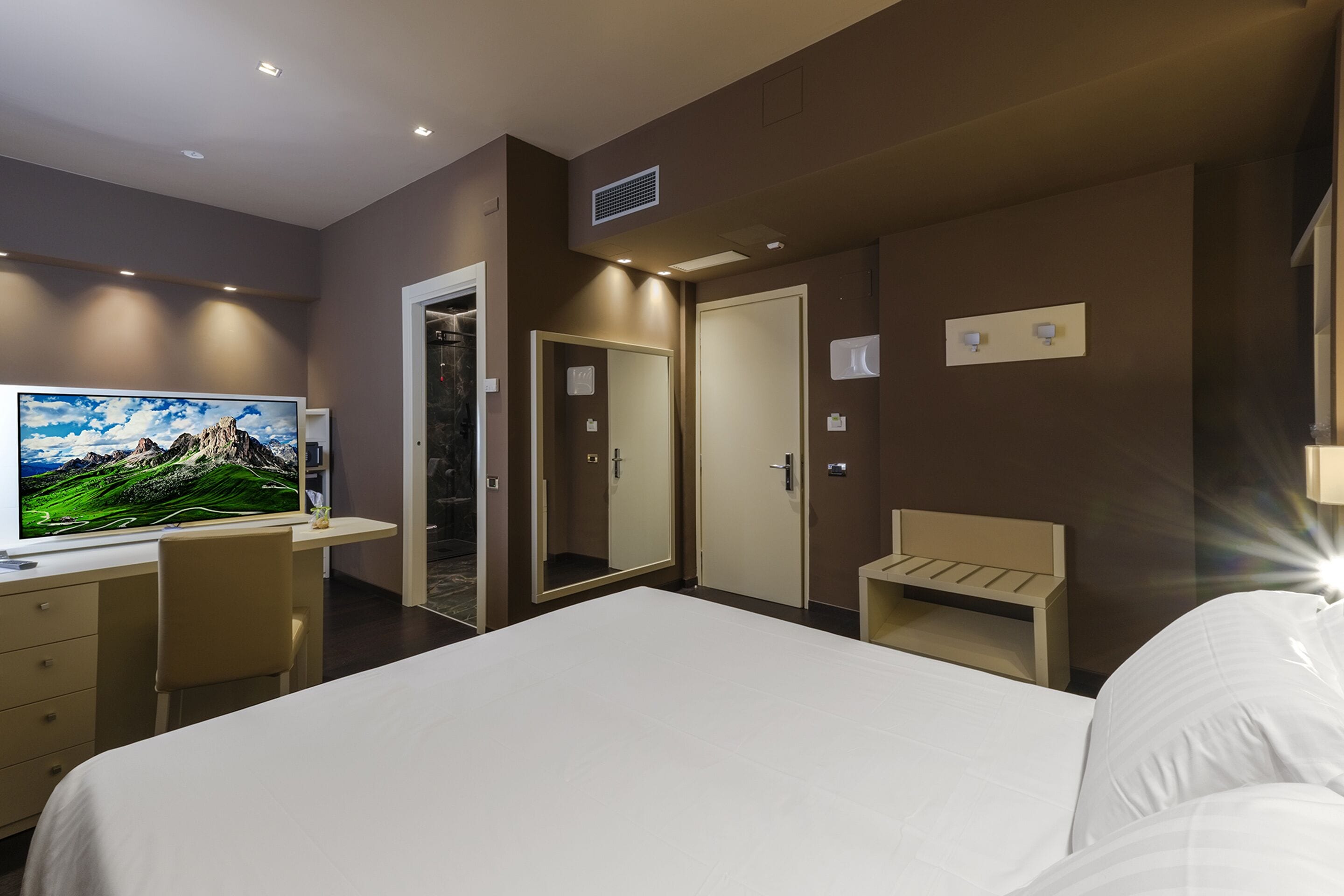 Ein modernes Hotelzimmer mit einem großen Bett, einem Schreibtisch mit Computer, einem Fernseher und einem Spiegel.