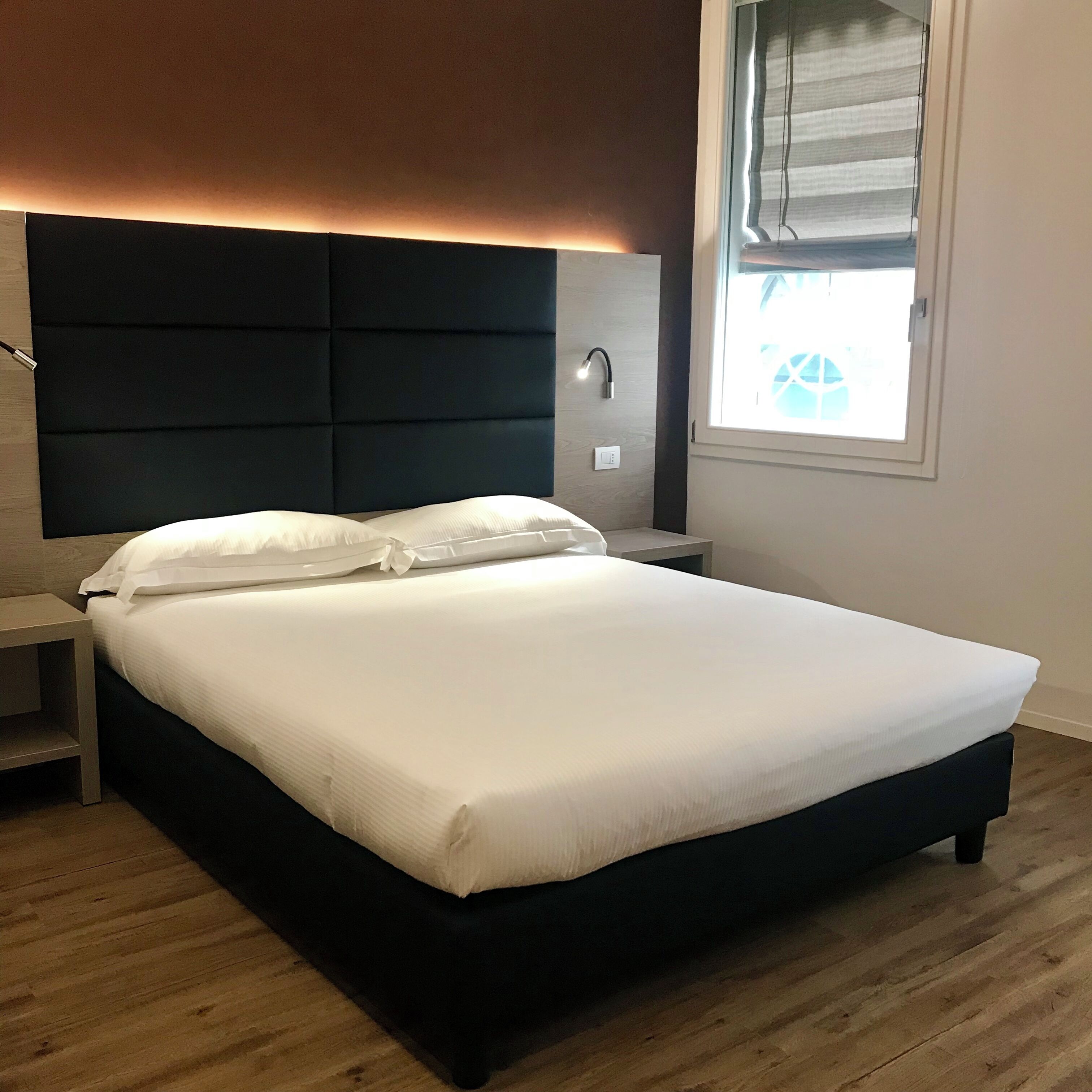 Un dormitorio con una cama grande, una mesita de noche, una lámpara de lectura fijada a la pared y una ventana con persianas.