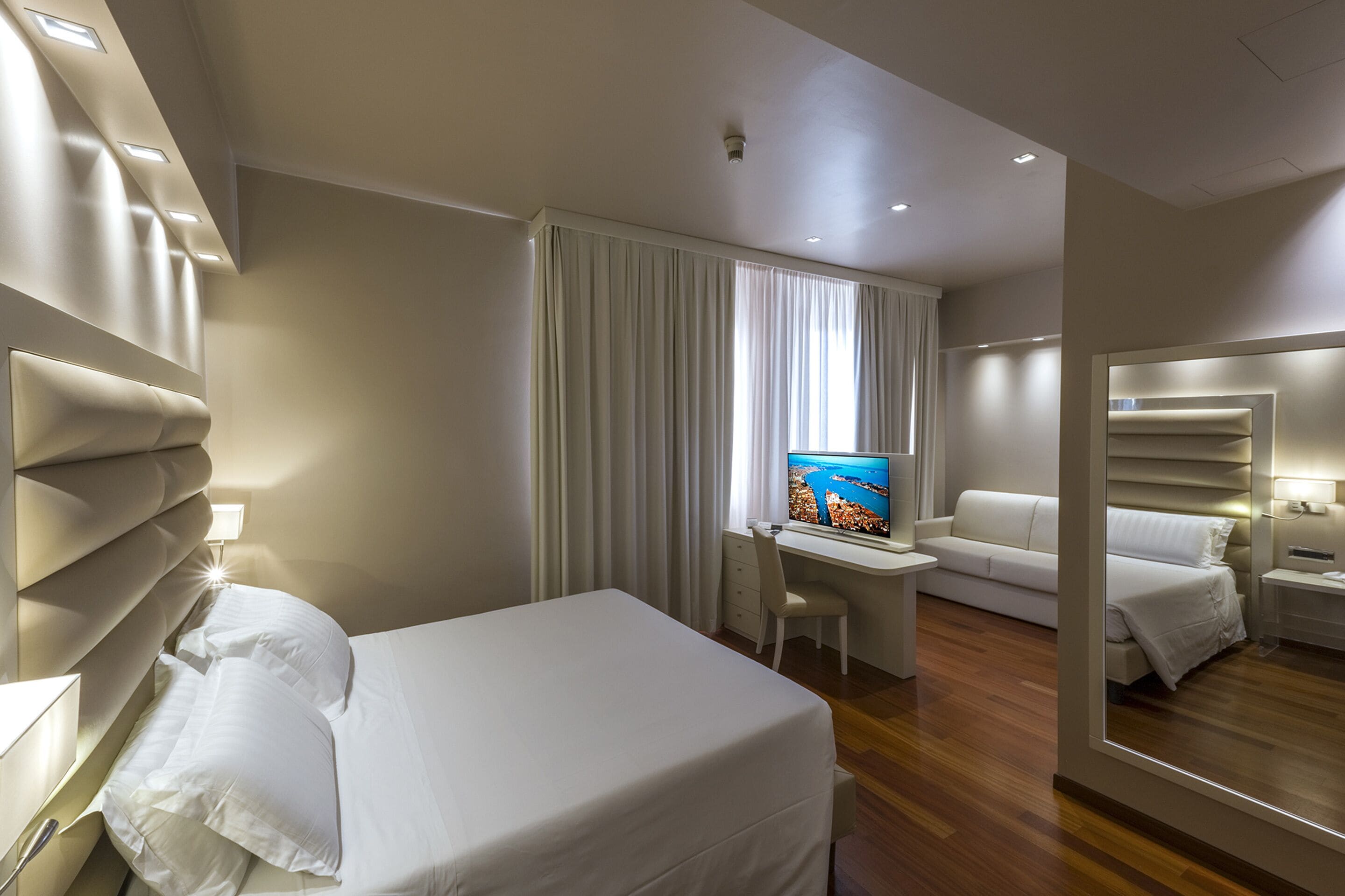 A hotel room with a bed, a desk with a TV, a sofa, and a mirror.
