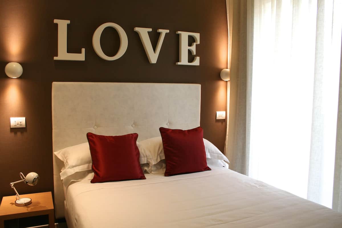 Habitación de hotel con una cama grande, dos almohadas rojas, cabecera blanca y una pared con la palabra 'LOVE' sobre la cama.