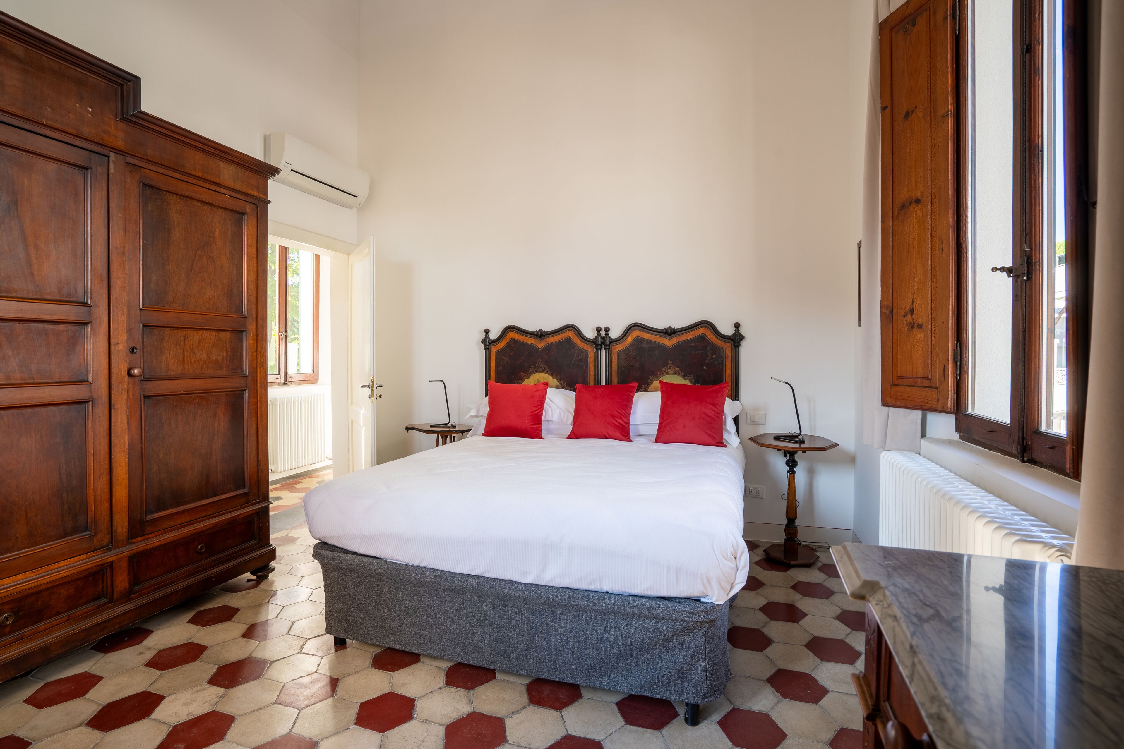 A bedroom with a large wooden wardrobe, a bed with red and white bedding, and a tiled floor.