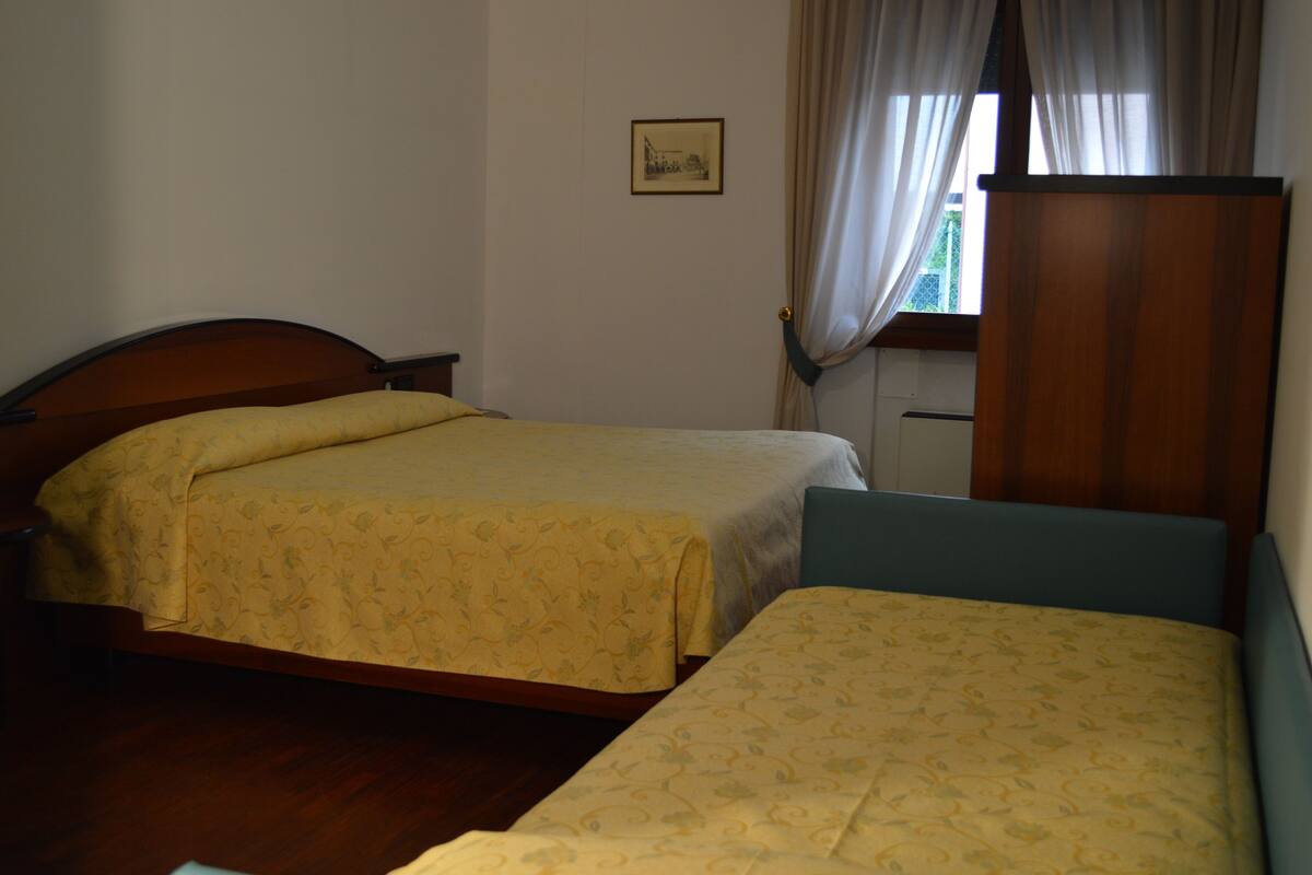 Une chambre avec deux lits, une tête de lit en bois, un petit bureau et une fenêtre avec des rideaux.