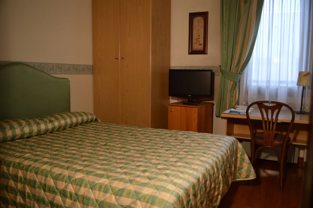 Une chambre d’hôtel comprenant un lit, un bureau avec une chaise, une télévision et une fenêtre avec des rideaux.