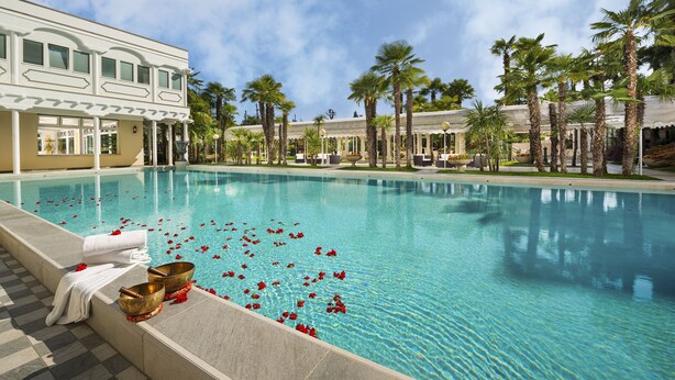 A swimming pool with a clear blue water, surrounded by palm trees and a building with a white facade. - HOTEL TERME METROPOLE – ORIENTAL SPA & RESORT (Abano Terme)