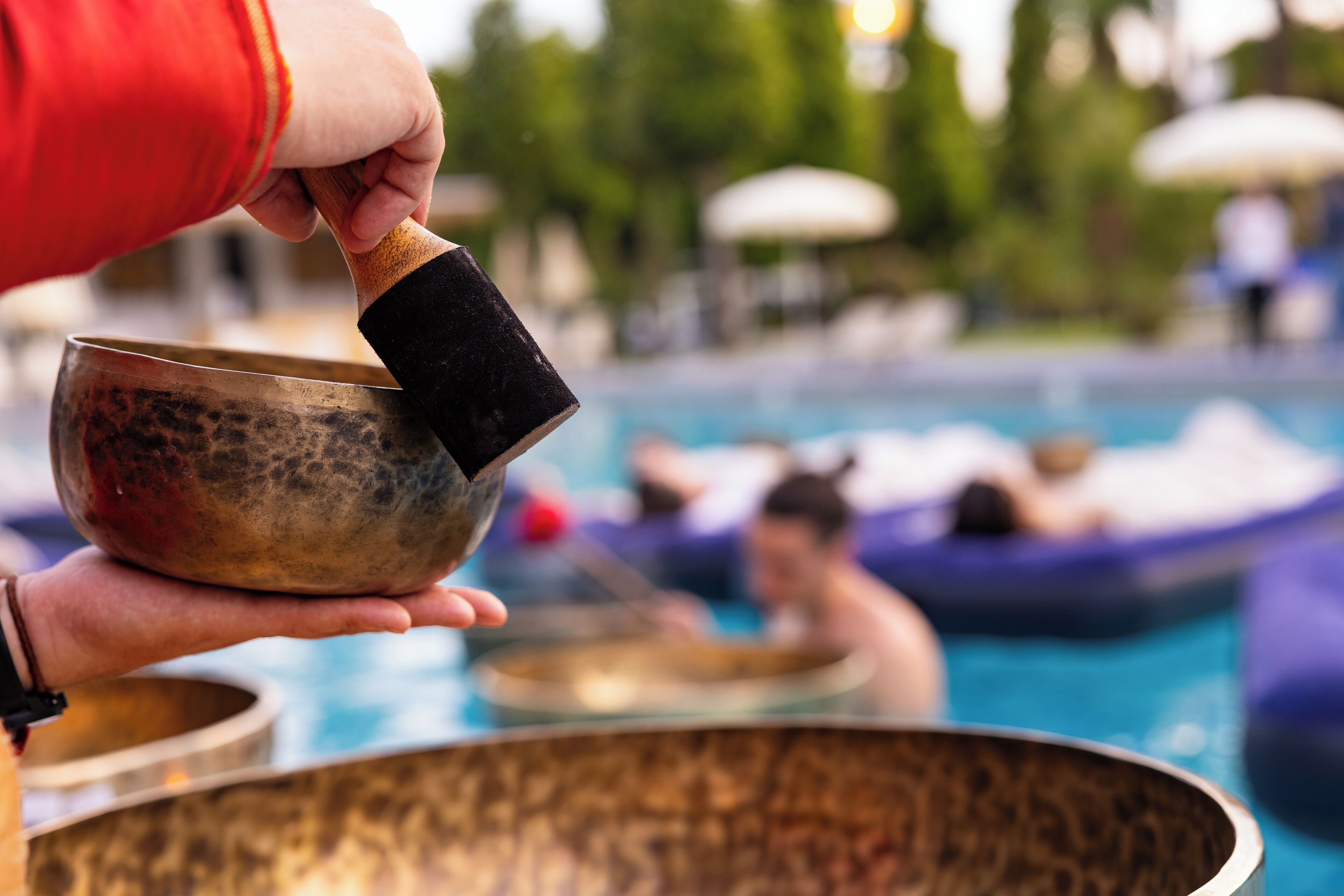 Una persona sostiene un cuenco metálico grande, probablemente usado para meditación o terapia de sonido, con una piscina y gente de fondo.
