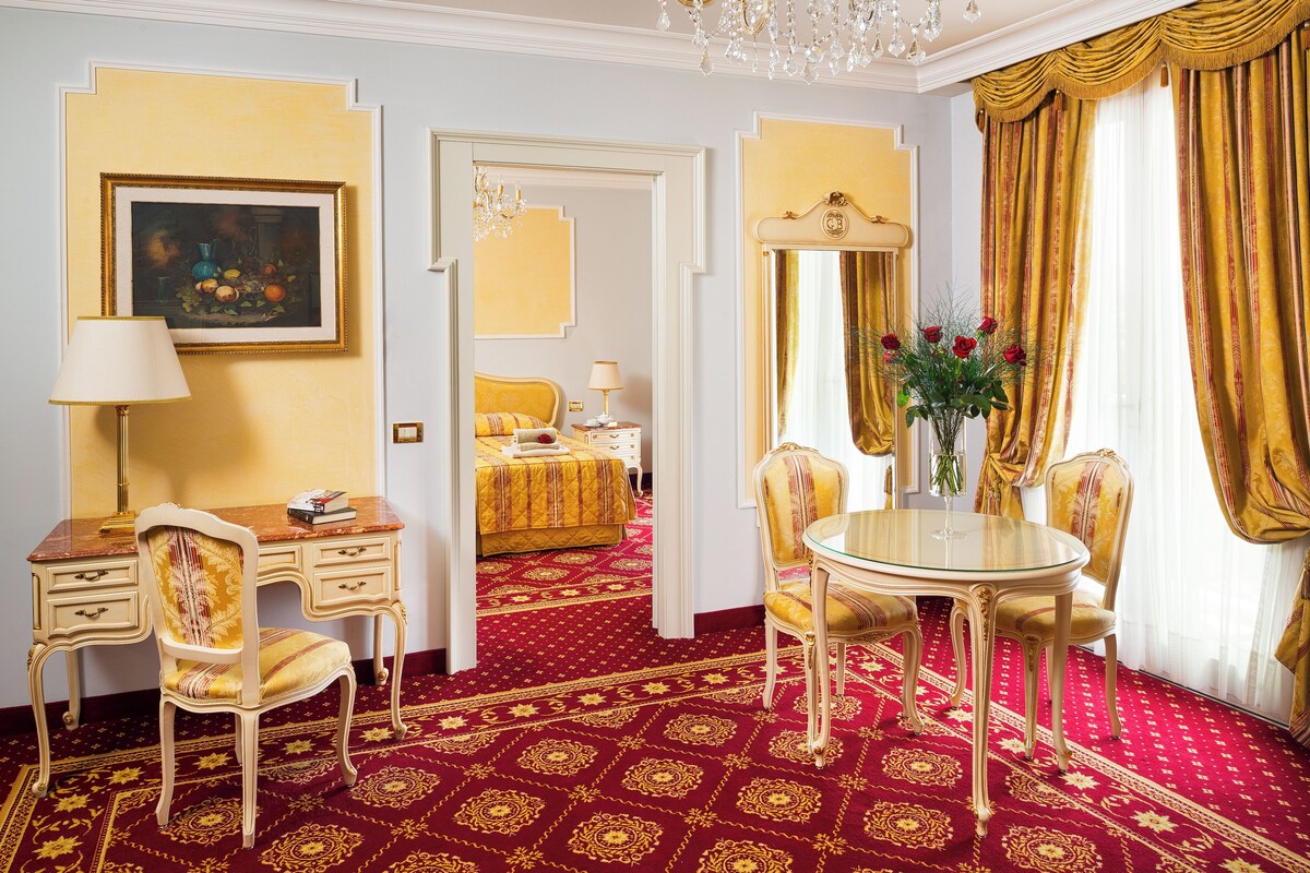 Habitación de hotel con alfombra roja, un escritorio con lámpara, una silla, una mesa redonda con sillas, una cama con cubrecama dorado y un cuadro en la pared.