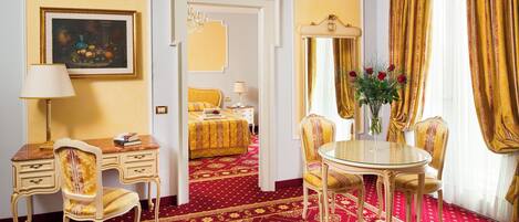 Habitación de hotel con alfombra roja, un escritorio con lámpara, una silla, una mesa redonda con sillas, una cama con cubrecama dorado y un cuadro en la pared.