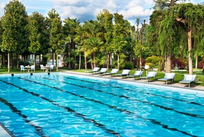 Una piscina con varias pistas, rodeada de tumbonas y árboles.