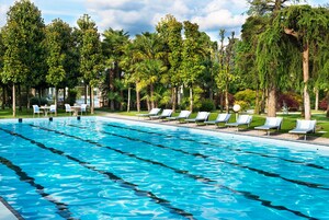 Una piscina con varias pistas, rodeada de tumbonas y árboles.