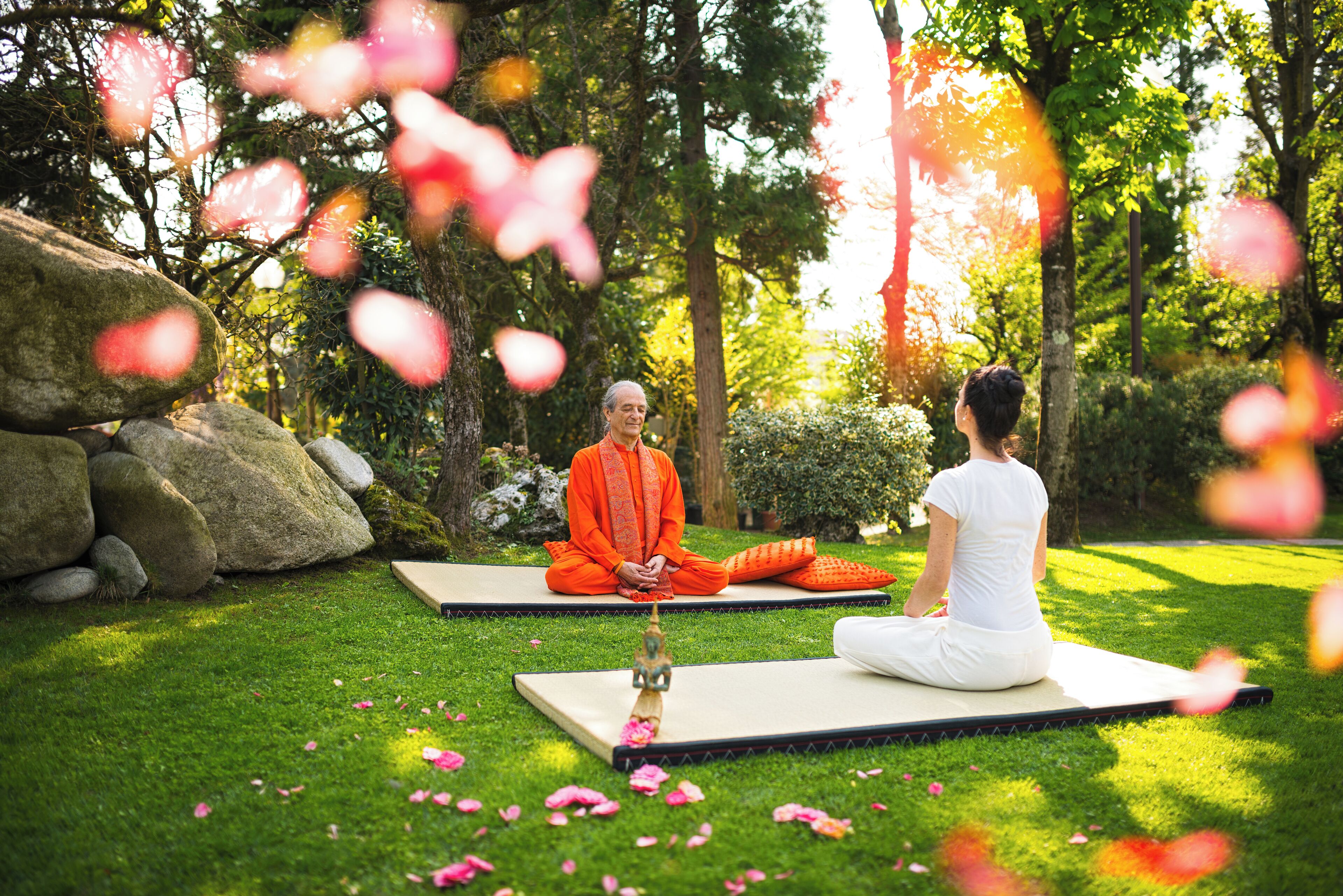 Two individuals meditating on mats in a garden with rocks and greenery.