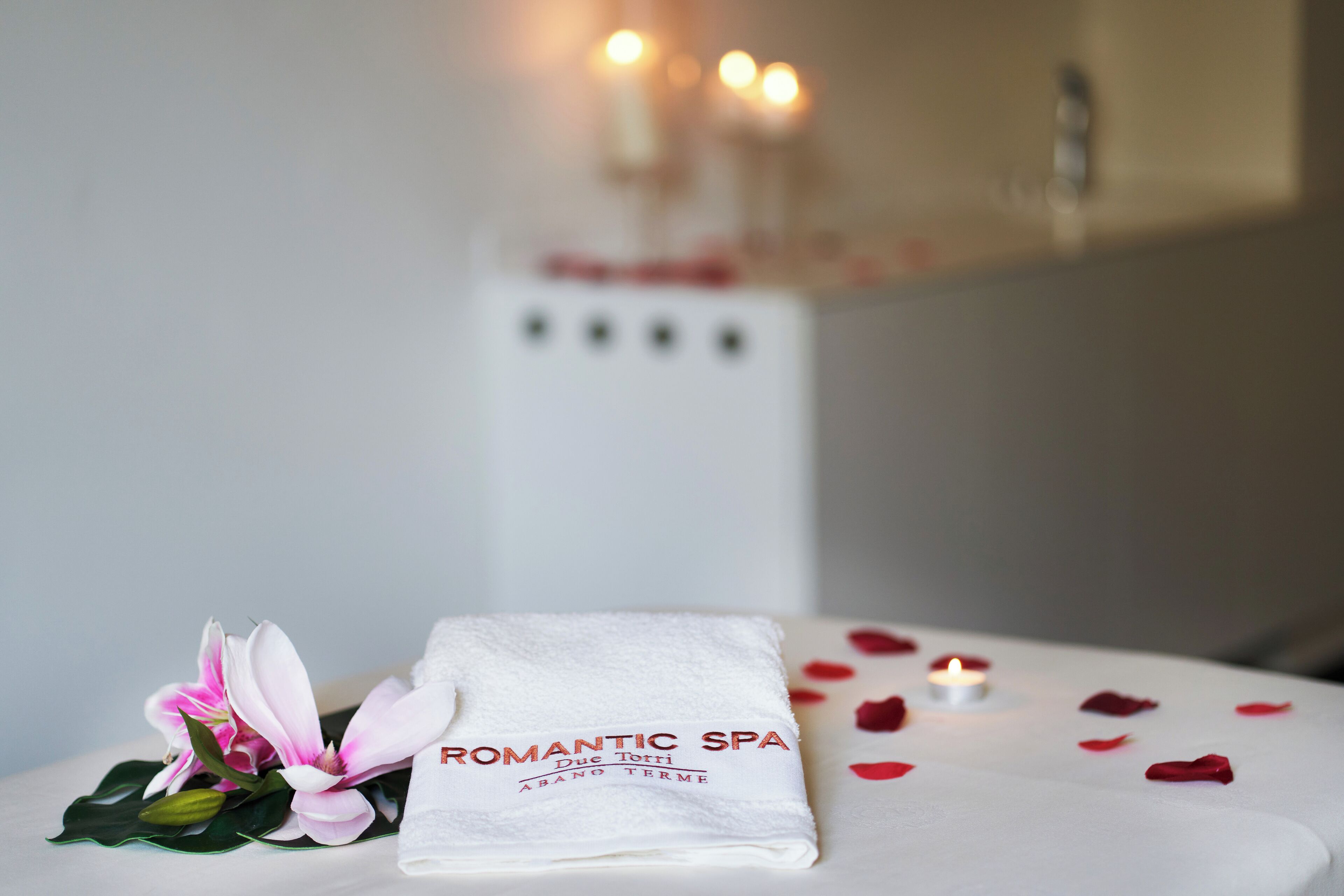 Una toalla blanca doblada con el logotipo 'Romantic Spa', una flor rosa y pétalos de rosa dispersos sobre una mesa.