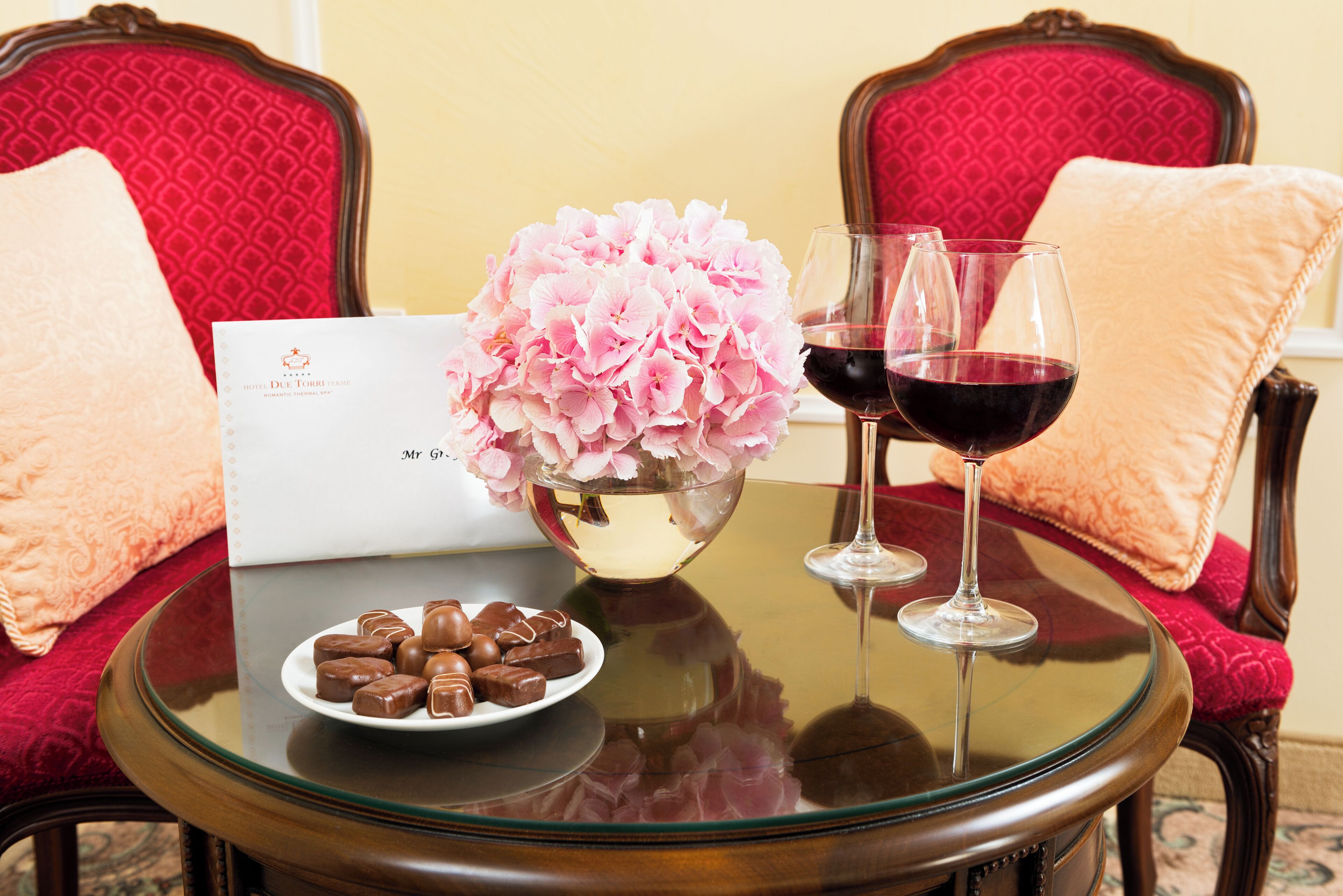 Un tavolo con un vaso di fiori rosa, due bicchieri da vino con vino rosso, un piatto di cioccolatini e una busta bianca.