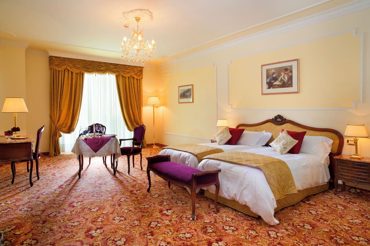 A hotel room with a large bed, two bedside tables, a desk with a chair, a chandelier, and a large window with curtains.
