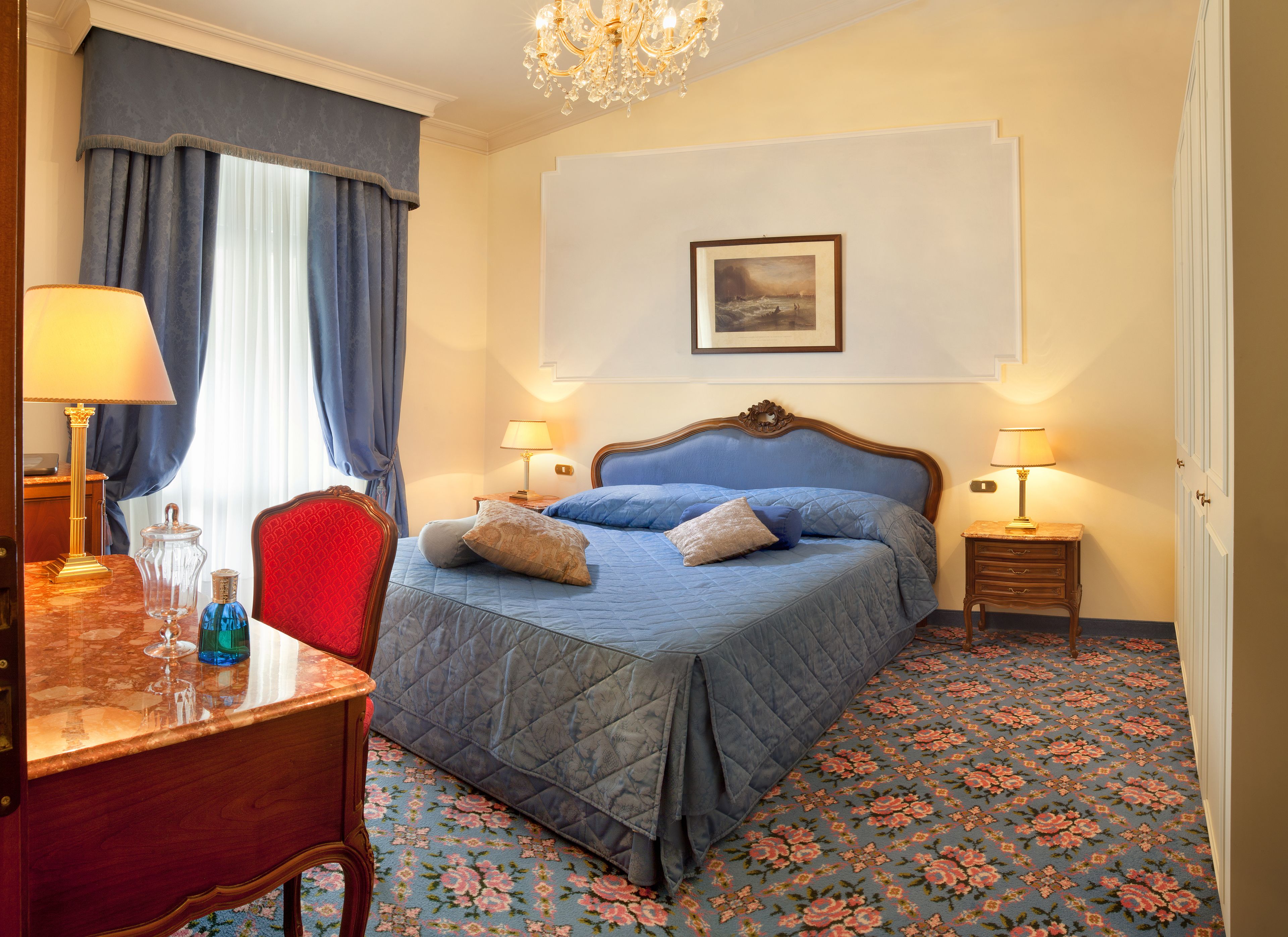 A hotel room with a bed, two bedside tables with lamps, a desk with a lamp, a chair, and a carpeted floor.