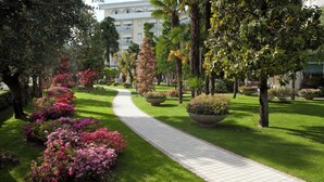 A landscaped garden with a paved walkway, palm trees, and flowering shrubs. -  HOTEL TERME DUE TORRI - Adults Hideaway & SPA (Abano Terme)