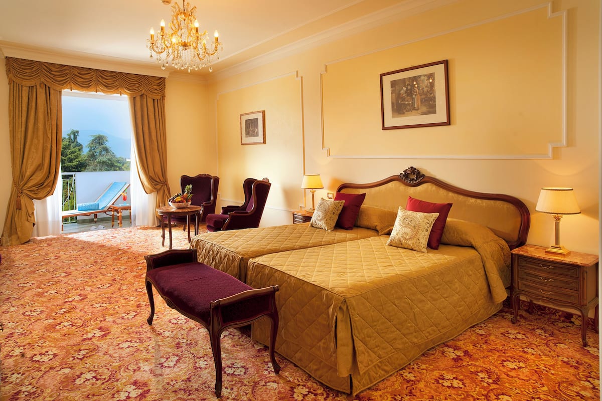 A hotel room with two beds, a sitting area with a chair and a table, a chandelier, and a balcony with a pool.