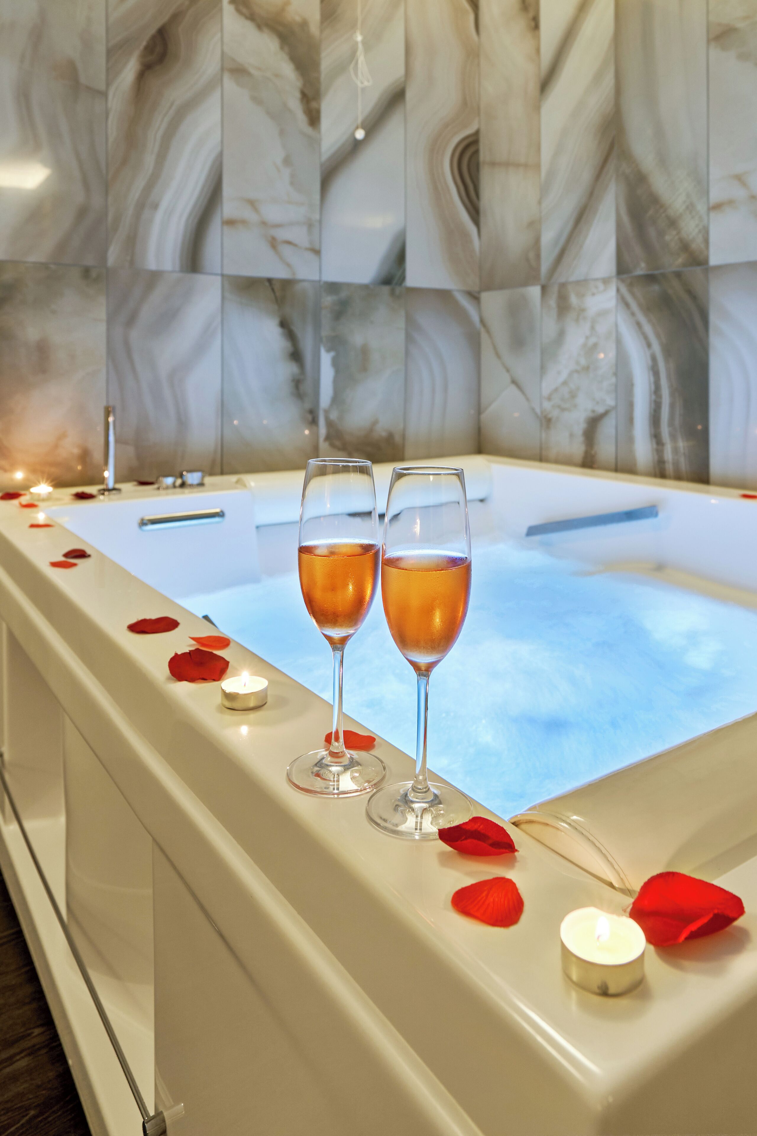 Une baignoire remplie de pétales de roses et entourée de bougies, deux verres de vin et, en arrière-plan, un mur de marbre.