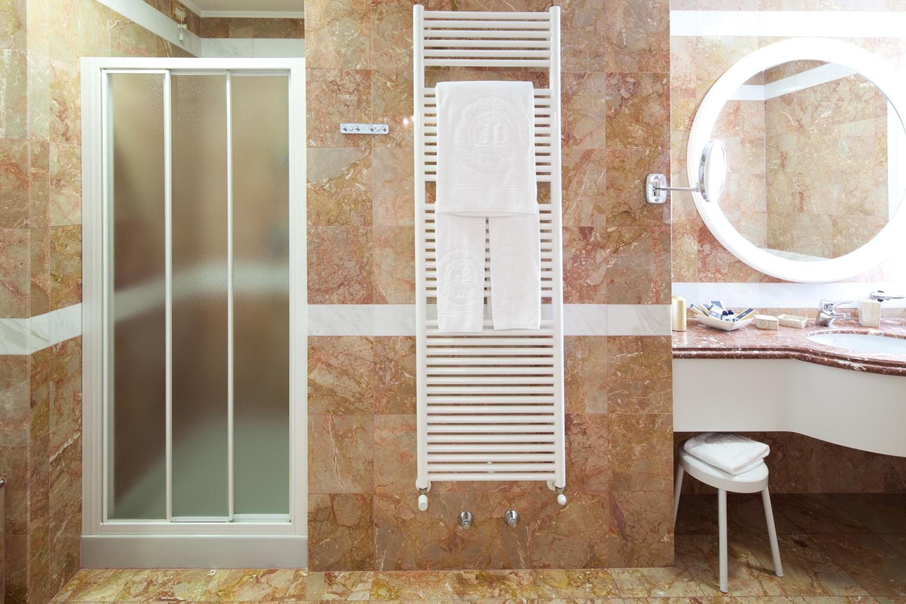 Un baño con mampara de vidrio, un perchero con toallas, un espejo redondo y un lavabo con artículos de aseo.