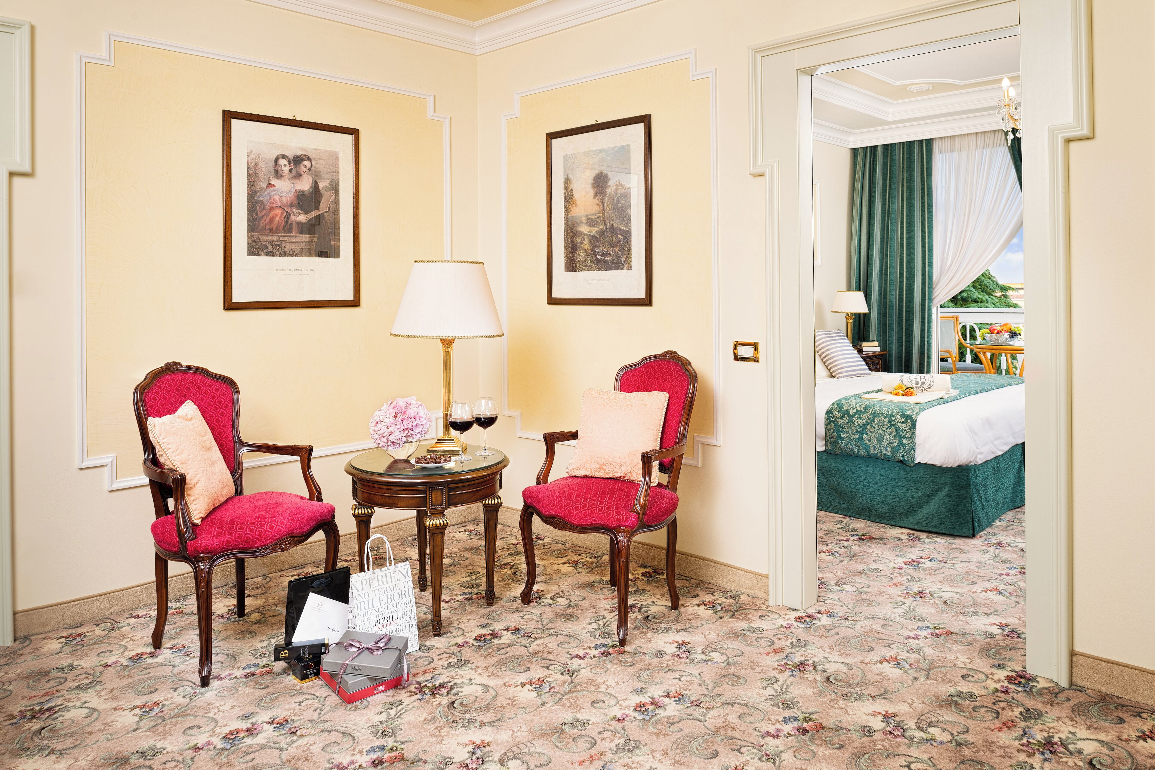 Habitación de hotel con dos sillones tapizados en rojo, una mesita redonda con lámpara, cuadros en las paredes y una cama con colcha verde.