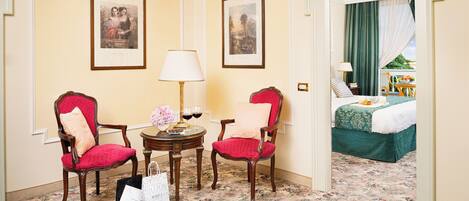 Habitación de hotel con dos sillones tapizados en rojo, una mesita redonda con lámpara, cuadros en las paredes y una cama con colcha verde.