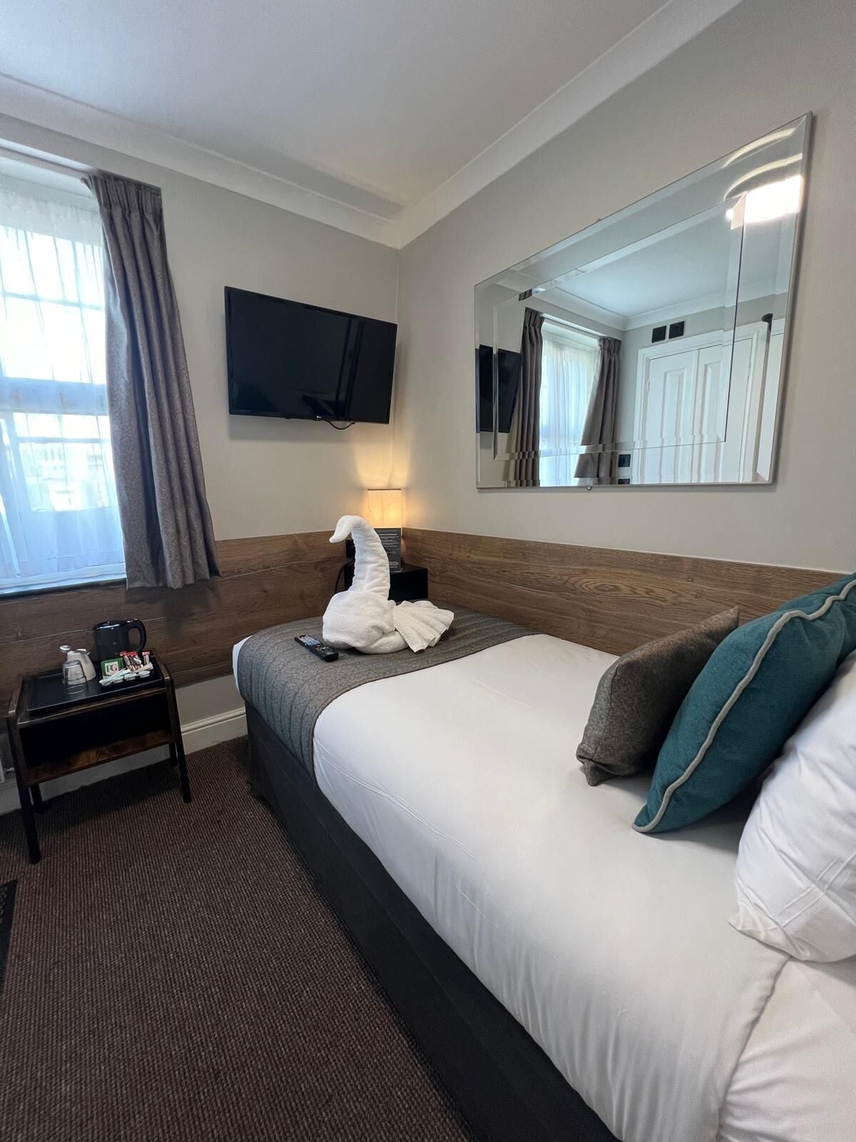 A hotel room with a bed, a flat-screen TV mounted on the wall, a bedside table with a lamp, and a window with curtains.