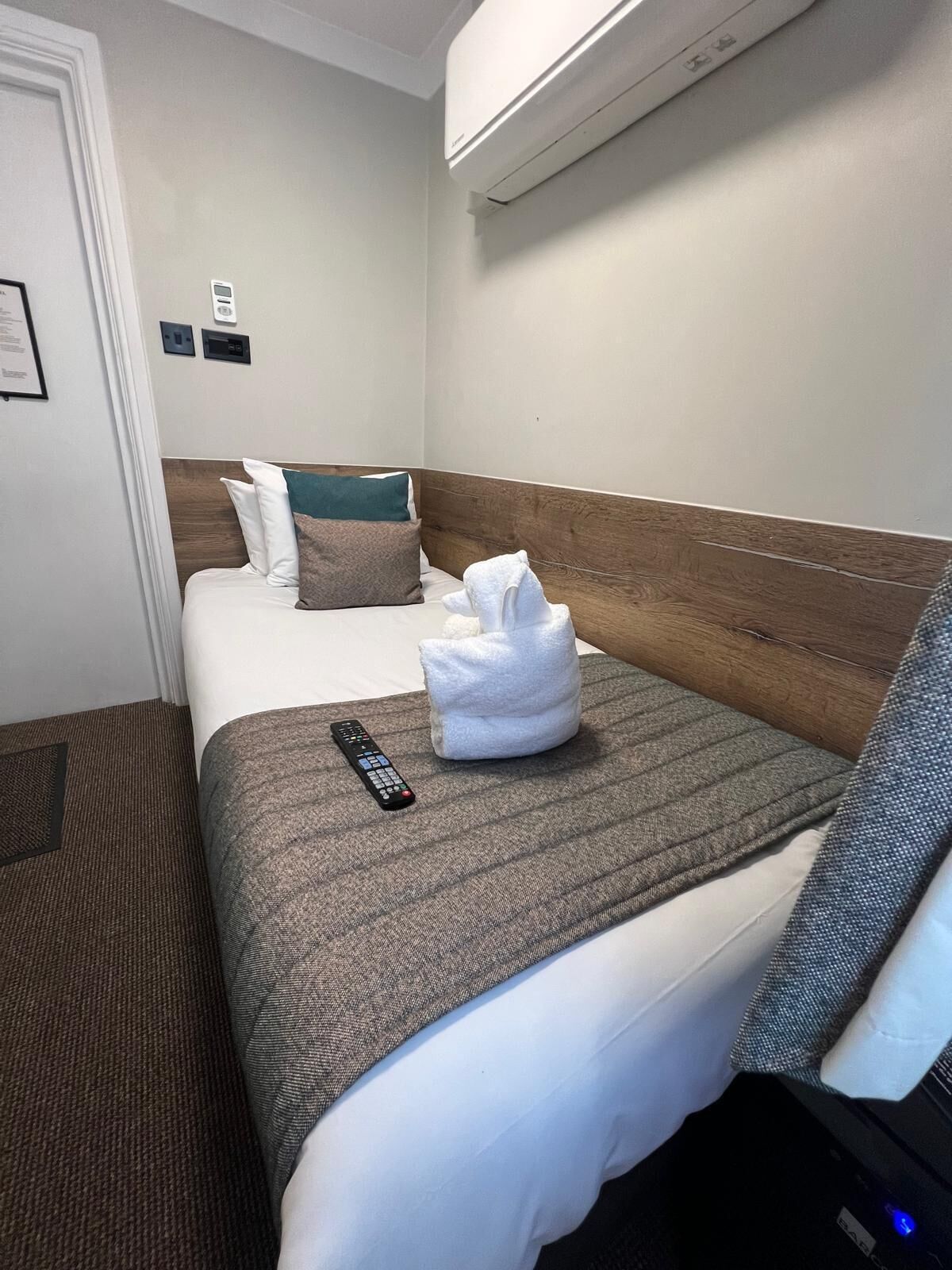 A hotel room with a bed, a remote control, a towel, and an air conditioning unit.