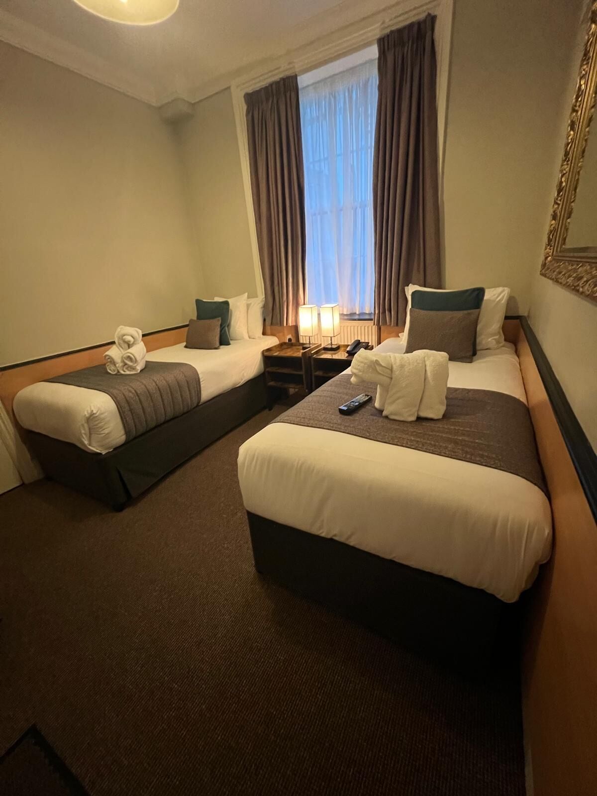 Quarto de hotel com duas camas, mesa de cabeceira com luminárias, janela com cortinas e espelho na parede.